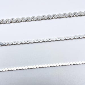 May include: Three silver-colored chain necklaces with a wavy, textured design. The chains are displayed against a white background, showcasing their delicate, interconnected links. The chains are likely for jewelry.