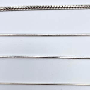 Puede incluir: Cuatro collares de cadena plateados de diferentes grosores se muestran sobre un fondo blanco. La cadena superior es la más gruesa, con un diseño de cadena de serpiente. Las otras tres cadenas son más finas, con un diseño de cadena de serpiente similar.