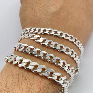 Könnte beinhalten: Nahaufnahme mehrerer silberner Panzerkettenarmbänder. Die Armbänder werden an einem Handgelenk getragen und haben eine glänzende, reflektierende Oberfläche. Die Ketten sind dick und haben ein klassisches, ineinandergreifendes Design.