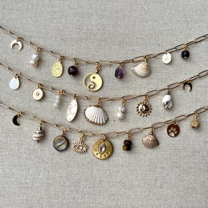 May include: Three gold chain necklaces with various charms, including seashells, a crescent moon, a yin yang symbol, a sun, a paw print, an evil eye, and a crystal.