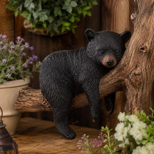 May include: A black bear cub figurine is resting on a tree branch. The bear has its eyes closed and is leaning against the tree. The figurine is surrounded by greenery and flowers, creating a naturalistic scene.