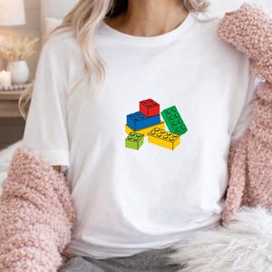 May include: A white short-sleeved t-shirt featuring a graphic print of a small pile of colorful plastic building blocks. The design includes individual blocks in solid red, blue, yellow, and green, arranged in a cluster on the center of the chest.