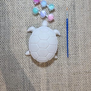 Puede incluir: Un kit de pintura de tortugas de cerámica. El kit incluye una figura de tortuga blanca, seis pequeños botes de pintura en varios colores pastel y un pincel azul fino. Los artículos están dispuestos sobre una superficie texturizada de tono neutro.