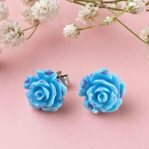 May include: A pair of stud earrings shaped like light blue roses with subtle iridescent purple accents on the petals. The earrings feature a metallic post backing and are displayed against a soft pink background with white flowers.