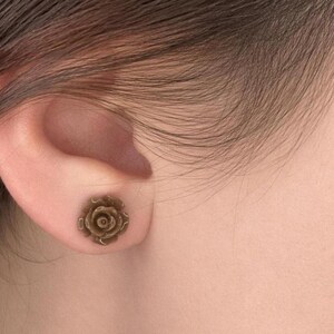 Könnte beinhalten: Ein kleiner, runder Ohrstecker in Form einer blühenden Rose. Der Ohrstecker hat ein metallisches, bronzefarbenes Finish mit komplexen, geschichteten Blütendetails, die eine texturierte und dreidimensionale Optik auf der Oberfläche erzeugen.
