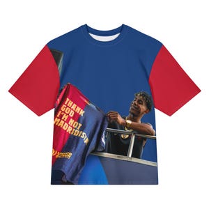 Può includere: Una maglietta blu, rossa e bianca con una persona che tiene in mano una maglia con la scritta "THANK GOD I'M NOT MADRIDISTA". La maglietta ha maniche rosse e un corpo blu. La maglia ha strisce rosse e blu.