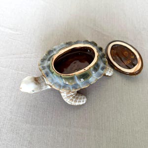 May include: A ceramic turtle-shaped trinket box with a removable lid. The shell features a glossy green and blue glaze, while the head and flippers are a light cream color with a grid pattern. The interior of the box is a deep, dark brown.