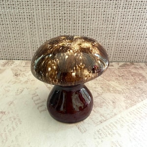 May include: A ceramic mushroom-shaped shaker with a glossy, dark brown cap featuring white, mottled, and speckled patterns. The base is a solid, smooth, dark brown color. The top of the cap has several small holes for dispensing contents.