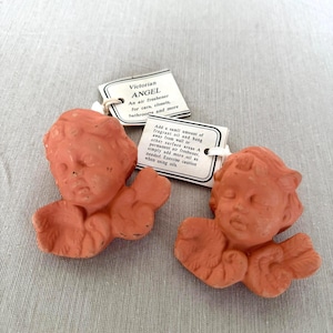 Puede incluir: Dos ambientadores con forma de ángel de color terracota con etiquetas blancas adjuntas. Las etiquetas incluyen el texto "Victorian ANGEL" e instrucciones para su uso con aceite aromático. Las figuras representan rostros de querubines con alas.