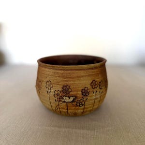 May include: A handcrafted, round ceramic bowl with a light brown exterior and a darker brown interior. The bowl features a delicate etched design of flowers and a bird. The bowl is suitable for decorative or functional use.