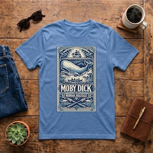 May include: Light blue t-shirt featuring a detailed graphic from the novel "Moby Dick" by Herman Melville. The design includes a whale, ship, and text. The shirt is displayed on a wooden surface with accessories.