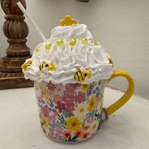 May include: A decorative floral mug with a yellow handle, topped with white whipped cream, yellow bee and cloud accents, and a straw. The mug features a colorful floral pattern with pink, yellow, and purple flowers.