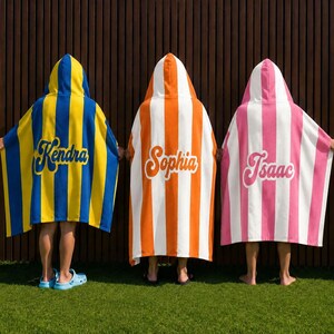 May include: Three hooded beach towels with personalised names. One is yellow and blue striped with "Kendra" in blue script. Another is orange and white striped with "Sophia" in white script. The third is pink and white striped with "Isaac" in pink script.