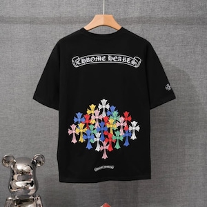 May include: A black short-sleeve t-shirt featuring the white text Chrome Hearts in a gothic font at the top. Below the text is a cluster of multi-colored crosses in shades of blue, red, yellow, green, and white. A small cross is on the sleeve.