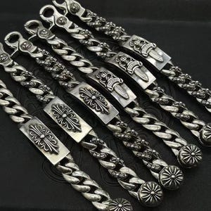 May include: Silver chain bracelets with decorative rectangular centrepieces. The bracelets feature a thick curb chain design and various embellishments, including crosses and dagger motifs. The bracelets are displayed on a dark surface.