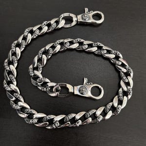 Pode incluir: Uma corrente de metal prateado com elos grossos e entrelaçados, apresentando motivos de cruz gravados. A corrente é fixada em ambas as extremidades por fechos de mosquetão funcionais, cada um detalhado com um pequeno emblema circular com uma cruz.
