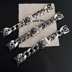 Puede incluir: Tres pulseras de cadena plateadas con diseños de cruces ornamentados. Cada pulsera presenta una gruesa cadena de eslabones y un cierre rectangular con un prominente motivo de cruz. Las pulseras se exhiben sobre una superficie oscura con un papel "Thank You".