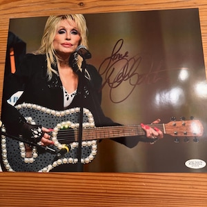 Può includere: Una fotografia lucida di Dolly Parton che si esibisce con un microfono e una chitarra decorata con perle bianche. L'immagine presenta uno sfondo scuro, una firma a inchiostro nero in alto a destra e un adesivo Total Sports Authentication.