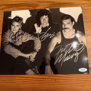 Può includere: Una fotografia in bianco e nero della band Queen con Roger Taylor, Brian May e Freddie Mercury. L'immagine include tre firme distinte sui soggetti e un adesivo Total Sports Authentics nell'angolo in basso a destra.