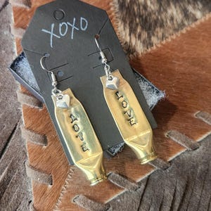 May include: A pair of handmade earrings made from brass bullet casings. Each earring has the word "LOVE" stamped on it, with a small heart charm and a silver hook. The earrings are displayed on a black card with "xoxo" written on it.