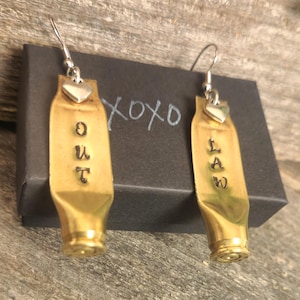 May include: A pair of brass-colored earrings with the word "OUT" stamped on one and "LAW" on the other. Each earring has a silver heart charm and a silver hook. The earrings are displayed next to a black box with the word "xoxo" written on it.