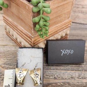 May include: A pair of earrings with a key and lock design. The earrings have a gold-colored base with a silver-colored key and lock charm. The earrings are displayed in a black box with the word "xoxo" written on it. A wooden box with succulents is in the background.