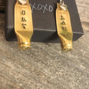 May include: A pair of brass-colored earrings made from bullet casings. Each earring has the word "OUTLAW" stamped on it. The earrings have silver-colored heart-shaped accents and are displayed on a black box.