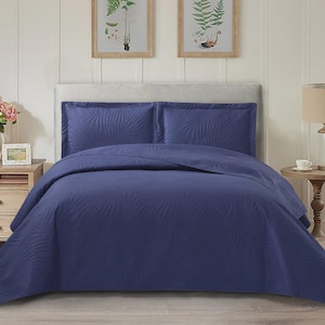 May include: A navy blue bedspread set with a textured, leaf-like pattern. The set includes a comforter and two pillow shams. The bedding is displayed on a bed with a light grey headboard, with a wooden nightstand and lamp on either side.