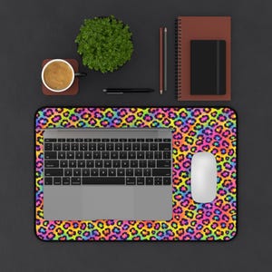 May include: A laptop, mouse, and keyboard sit on a vibrant, rainbow-colored leopard print mousepad. A cup of coffee, a small plant, a notebook, and writing utensils are also on the dark surface.