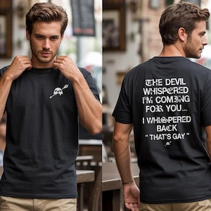 May include: A black short-sleeve t-shirt featuring a small white skull and arrow graphic on the front chest. The back displays the white text: THE DEVIL WHISPERED I'M COMING FOR YOU... I WHISPERED BACK "THAT'S GAY" with a small skull icon below.