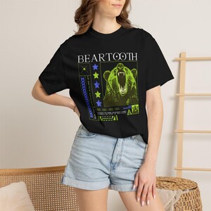 Puede incluir: Camiseta negra con un diseño gráfico de un oso rugiente en verde neón y blanco, y la palabra "BEARTOOTH". El diseño incluye estrellas azules y verdes y texto adicional. Una camiseta gráfica informal.