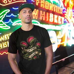 May include: A black short-sleeved t-shirt featuring a graphic print of several red and green Venus flytrap plants. A beige ribbon banner is wrapped around the plants with the text EAT THE RICH printed in black capital letters on the banner.