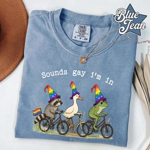 Könnte beinhalten: Hellblaues Comfort Colors T-Shirt mit dem Text "Sounds gay I'm in". Das Shirt zeigt einen Waschbären, eine Ente und einen Frosch auf Fahrrädern, jeder mit einem Regenbogen-Zaubererhut. Am Fahrrad des Waschbären ist eine Regenbogenflagge befestigt.