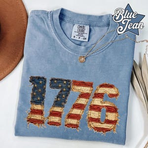 May include: A light blue t-shirt with the number "1776" in a distressed American flag design. The shirt has a crew neck and short sleeves. A gold necklace with a pendant is draped across the shirt. The words "Blue Jean" are in a star-shaped logo.