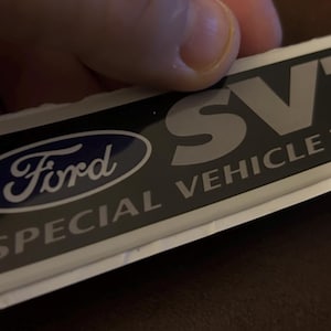 May include: Rectangular sticker featuring the Ford logo in blue and white, with the text "SVT SPECIAL VEHICLE TEAM" in white on a black background. The sticker is held by a hand.