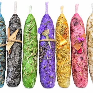 May include: Seven cylindrical wax candles in blue, black, green, purple, yellow, pink, and white. Each candle is textured with embedded dried herbs, flowers, or wood shavings, tied with twine, and features a small wick at the top centre.