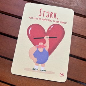 Könnte beinhalten: Rechteckige Karte mit dem Wort "Stark" in roter Schrift. Die Karte zeigt eine Cartoon-Illustration eines Kraftmanns, der eine Langhantel aus einem roten Herzen hebt. Der Kraftmann trägt ein blaues Tanktop und hat einen rosa Schnurrbart.