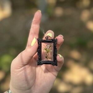 May include: A small, black framed pendant with a pressed flower inside. The pendant is rectangular with ornate detailing. The background is blurred, suggesting an outdoor setting. The pendant is held in a hand with yellow nail polish and a silver bracelet.