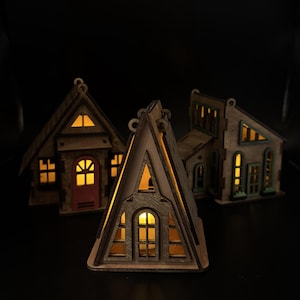 May include: Three miniature wooden house structures with laser-cut details and warm internal lighting. The front house is an A-frame design, while the two houses behind feature pitched roofs, windows, and doors, all glowing with a soft yellow light.