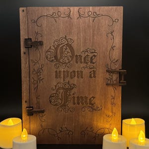 May include: A rectangular wooden book-shaped box with a dark brown finish, featuring the laser-engraved text "Once upon a Time" on the front cover, surrounded by a decorative vine border and secured with two dark metal hinges on the side.