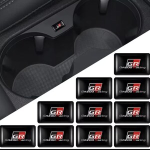May include: Black car interior featuring a cup holder and a collection of black rectangular stickers. Each sticker displays "GR" in white and red, with "GAZOO Racing" underneath.