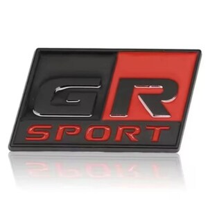 Peut inclure: Un emblème GR SPORT noir et rouge. L'emblème est rectangulaire avec une section supérieure gauche noire et une section supérieure droite rouge. Les lettres "GR" sont argentées et "SPORT" est en rouge.