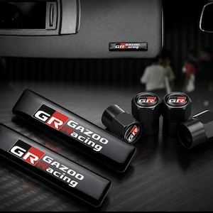 May include: Two rectangular black emblems and four black valve stem caps featuring the Gazoo Racing logo. Each piece displays the GR emblem in red, white, and black with the text Gazoo Racing. The items have a glossy finish and are arranged together.