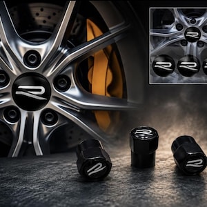 May include: A set of four black cylindrical tire valve caps featuring a white stylized letter R logo on the top. The caps are displayed alongside a car wheel with a matching R logo center cap and a gold brake caliper visible in the background.