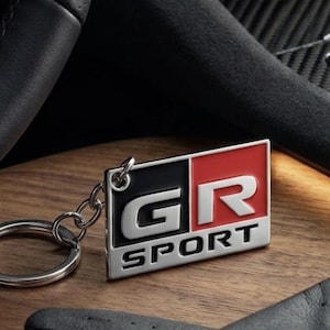 May include: A rectangular metal keyring featuring the GR SPORT logo with a black and red enamel finish. The design includes the letters GR in a bold silver font above the word SPORT in a smaller silver font, attached to a silver metal ring.