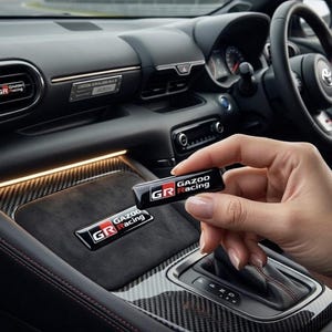 May include: A rectangular black emblem featuring the white and red Gazoo Racing logo with the text Gazoo Racing. The emblem is held in front of a car interior dashboard that features a matching emblem on a dark suede surface and carbon fibre trim.
