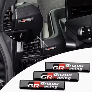 Puede incluir: Pegatinas negras y rojas con el emblema "GR Gazoo Racing" para coche. Las pegatinas son rectangulares con las letras "GR" en blanco y rojo, y las palabras "Gazoo Racing" en blanco. Las pegatinas se muestran en un salpicadero de coche.