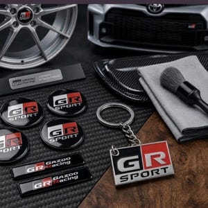 Puede incluir: Una colección de accesorios automotrices GR Sport que incluye cuatro emblemas circulares, dos insignias rectangulares Gazoo Racing, un llavero de metal, un modelo de rueda plateado, un cepillo de limpieza y un paño de microfibra, todos con el logotipo GR Sport.