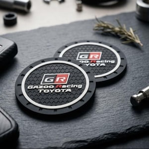 May include: Two circular black rubber coasters featuring a textured honeycomb pattern and a white border. The center displays the red, white, and black GR logo with the text Gazoo Racing Toyota printed in white lettering below the emblem.