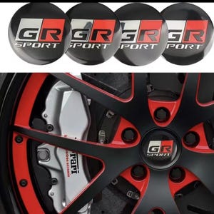 Può includere: Quattro coprimozzi neri con la scritta "GR SPORT" in bianco e rosso. L'immagine mostra anche un primo piano di una ruota di un'auto con raggi neri, dettagli rossi e una pinza freno argentata.